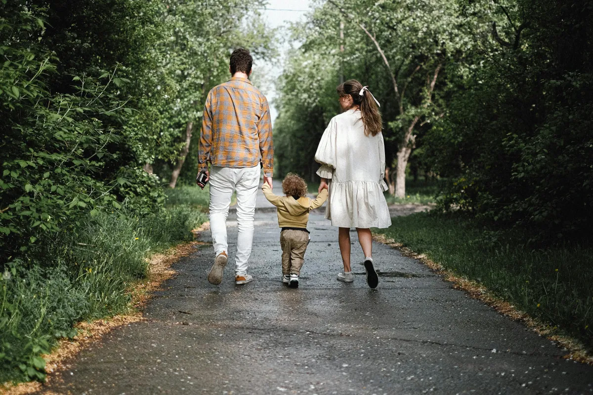 A joyful moment of a family walking together along a tree-lined path, with a child holding hands with both parents. The lush greenery and casual outdoor setting evoke the relaxed, inviting spirit of local parks and riverwalks.