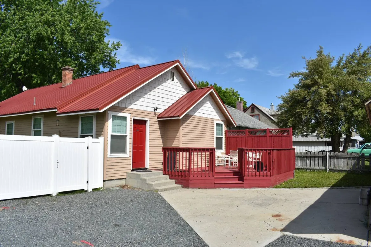 A welcoming red deck bordered by a white fence, surrounded by green trees and featuring a front door nearby. The space looks ideal for gatherings, relaxing outdoors, and enjoying the privacy of a fenced yard.