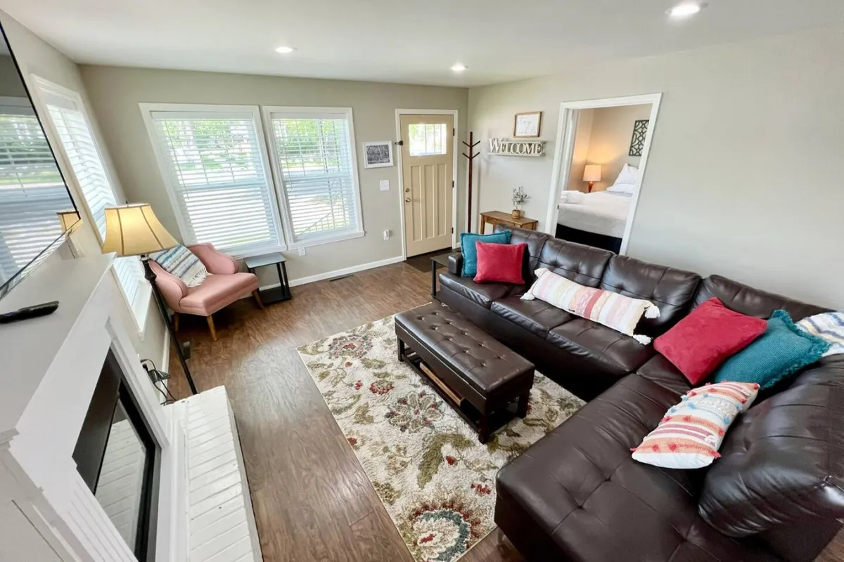 A warm and inviting living room with a leather sofa, colorful cushions, a decorative rug, a welcoming sign, and a cozy lamp. The space is arranged for both relaxation and group conversation, making it ideal for family and friends to gather.