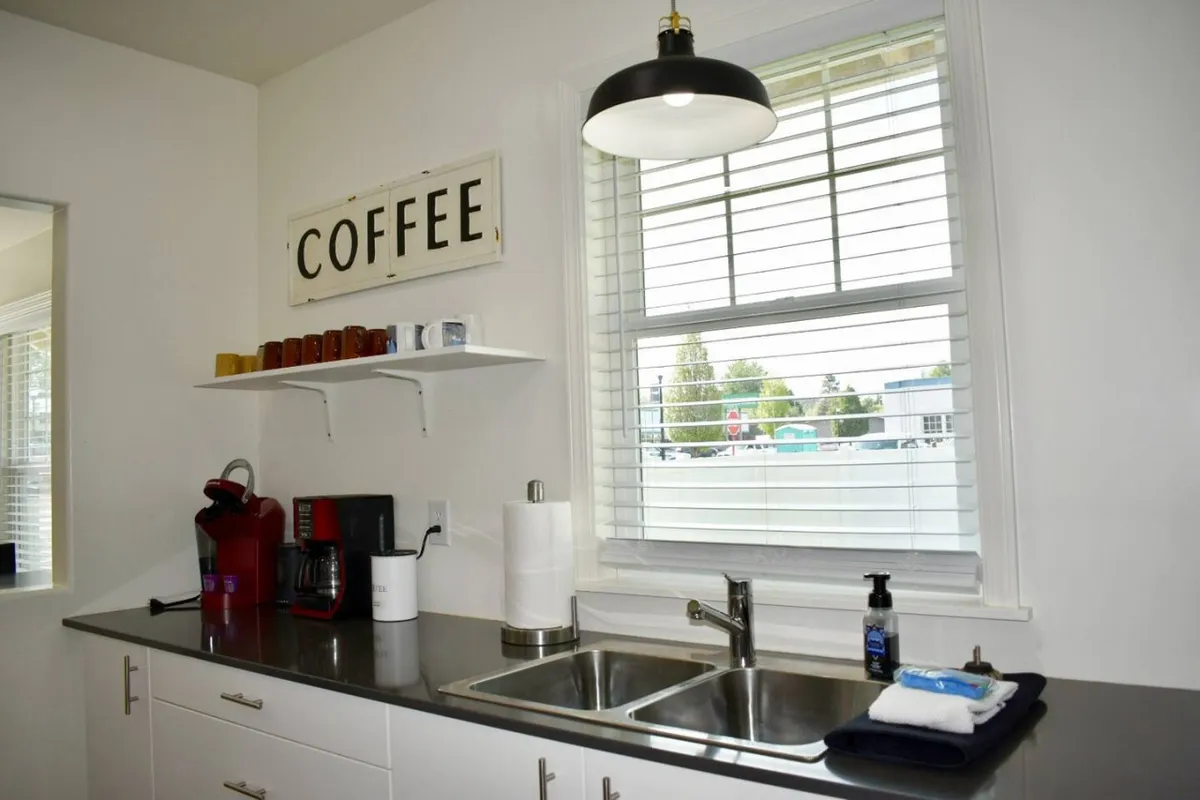 A close-up of a kitchen coffee station, including a coffee maker, sink, a shelf stocked with mugs, and a window letting in filtered natural light. The setup is warm and inviting, perfect for morning routines.