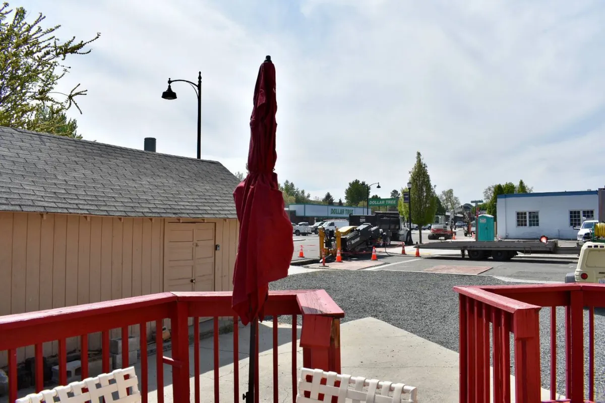 A cozy wooden deck with a red umbrella overlooks the neighborhood, surrounded by local buildings. The outdoor seating area invites guests to relax, sip coffee, or people-watch, emphasizing both leisure and engagement with the local scene.