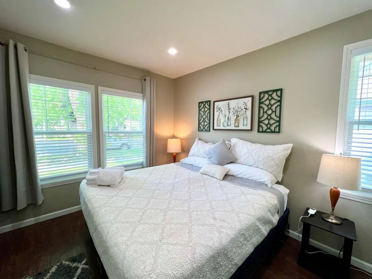A serene and comfortable bedroom featuring a bed with decorative pillows, a nightstand with a lamp, framed wall art, and large windows with curtains. Natural light streams in, creating a relaxing atmosphere ideal for restful nights.