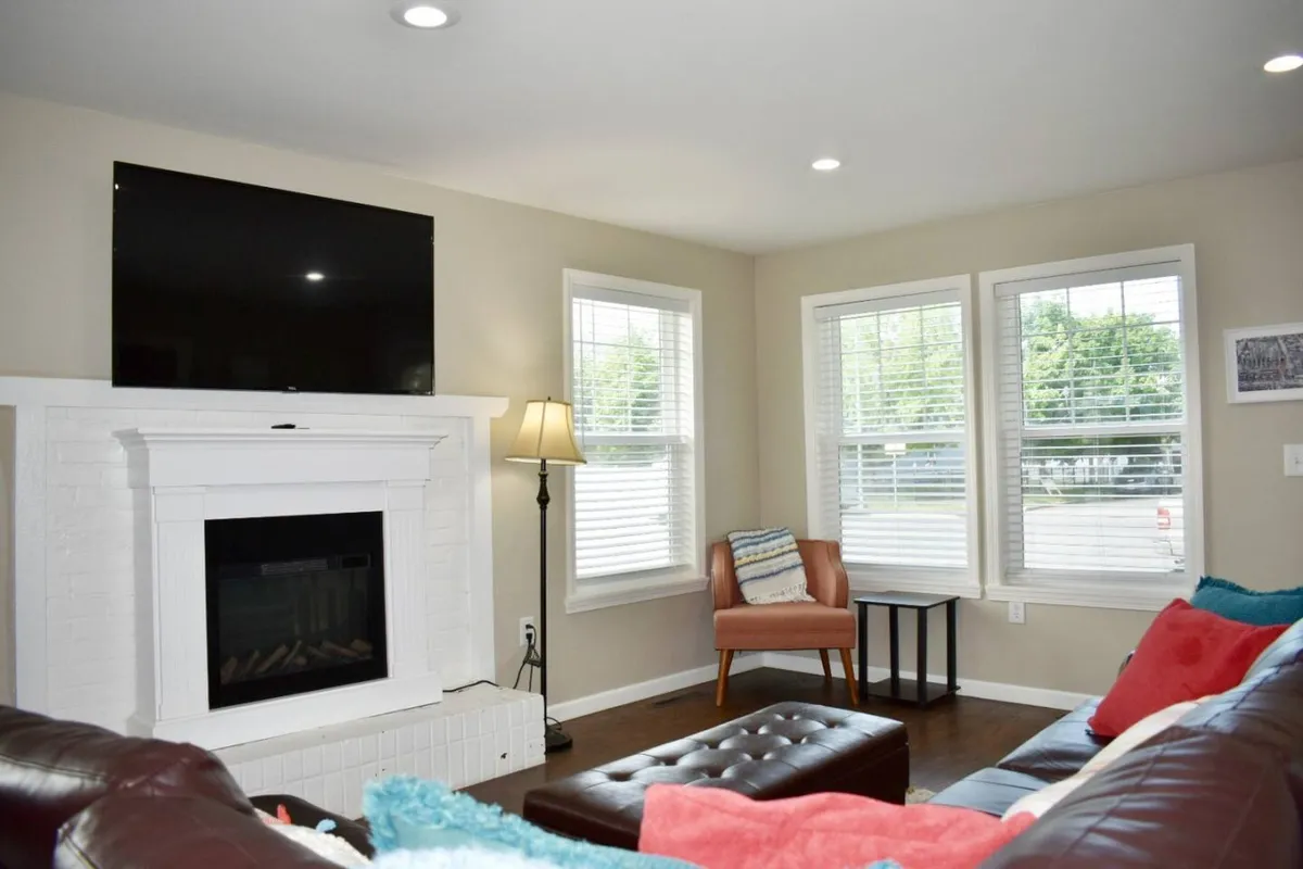 A cozy living room featuring a plush leather sectional, a wall-mounted television, a fireplace, comfortable seating, and large windows that allow plenty of natural light. The original wood floors add a sense of historic charm while the modern furnishings create a welcoming social atmosphere.
