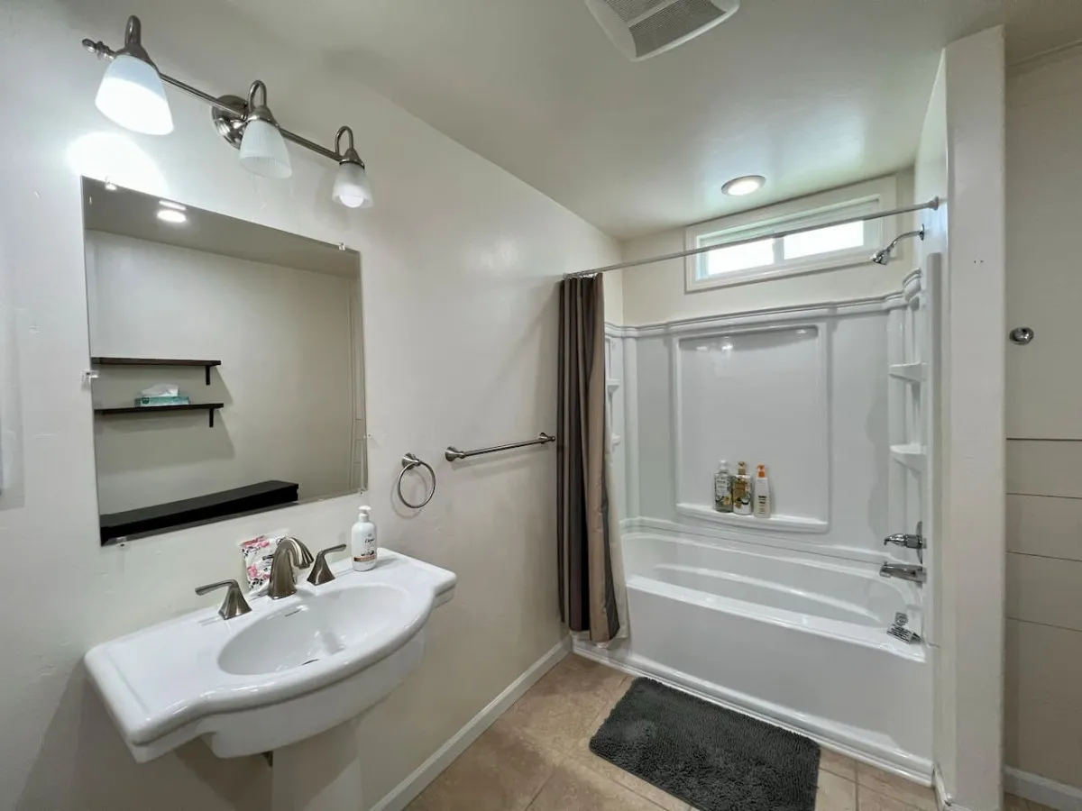 A bright and practical bathroom featuring a sink, shower area, mirror, and shelves. The remodeled space is inviting and functional, perfect for relaxing soaks or bathtime routines with children.