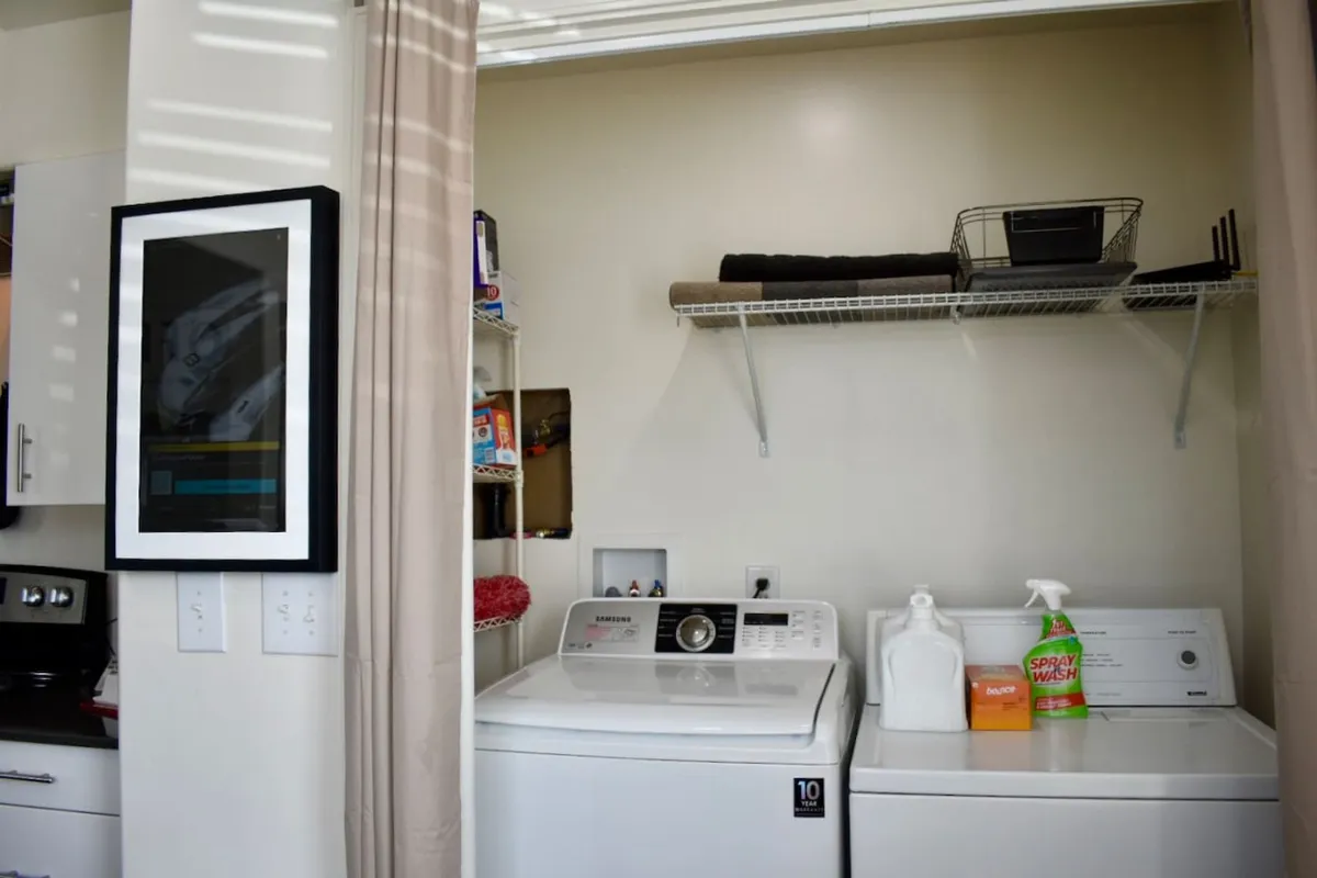 A practical laundry nook featuring a washing machine, dryer, organized shelf with laundry supplies, and framed art, tucked efficiently into the living space.