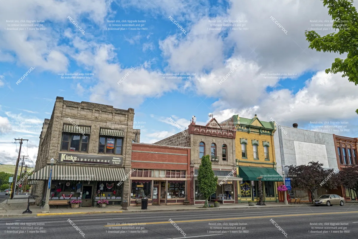 A vibrant exterior scene showing the Kicks Sportswear storefront along Main Street, with colorful Victorian architecture and trees lining the sidewalk. Shoppers and diners contribute to the lively, bustling atmosphere of Baker City's historic downtown.