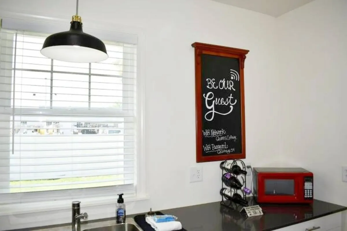 Inside, a friendly kitchen scene centers on a chalkboard with a welcoming 'Be Our Guest' message, a red microwave, and a sink area. The chalkboard hints at details like WiFi, crucial for remote workers, while the kitchen setup underscores the at-home comfort for extended stays and easy meal prep.